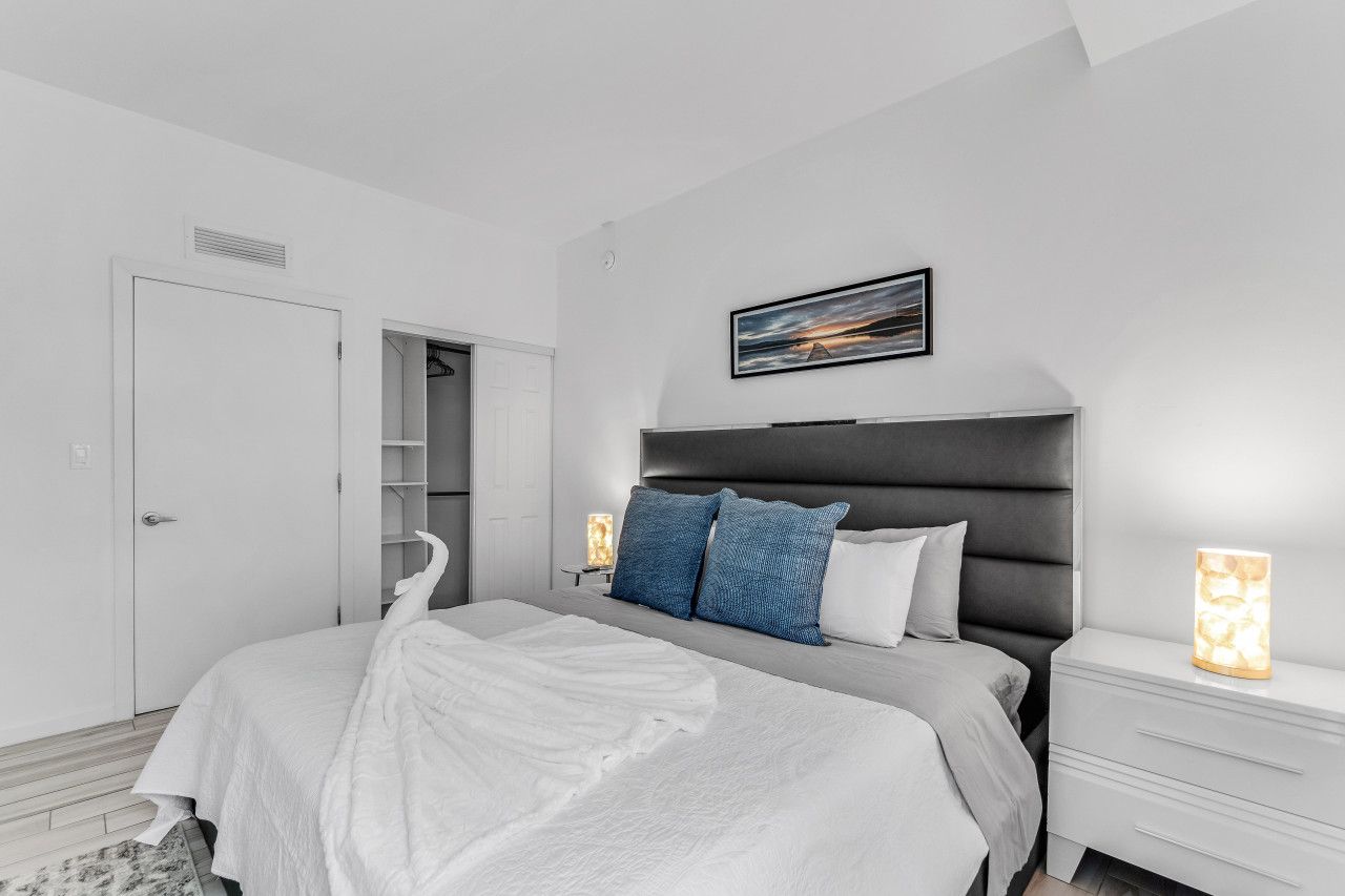 A modern bedroom with a large bed, a headboard, a nightstand, a bedside table, a wardrobe, and a framed picture on the wall.