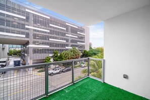 Een balkon met kunstmatig groen gras, een metalen hekwerk en uitzicht op een modern gebouw met horizontale lijnen en een parkeerplaats eronder.