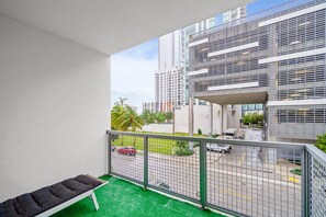 A balcony with a green carpet, a cushioned bench, and a metal railing. In the background, there are modern buildings and a road with vehicles. - Lovely Unit Mins from Wynwood with Free Parking (Miami)