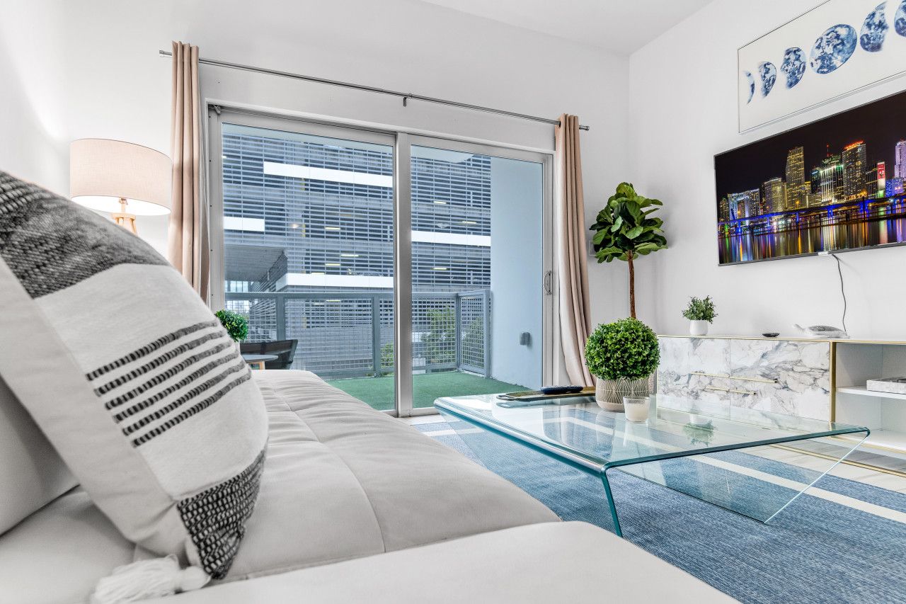 A modern living room with a sofa, a glass coffee table, a cityscape painting, and a balcony with a view of buildings.