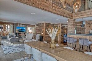 Una sala moderna con una pared de acento de madera, un televisor de pantalla plana grande, una zona de comedor con mesa de madera y taburetes de bar, y una sala de estar con sofá y sillones.