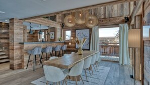 Een ruime eethoek met een lange houten tafel en moderne stoelen, een keuken met een bar, en een groot raam met uitzicht.
