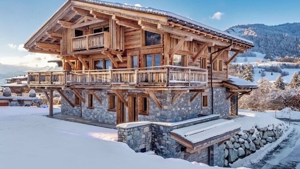 Een houten chalet met balkon, stenen fundering en een besneeerd landschap op de achtergrond.