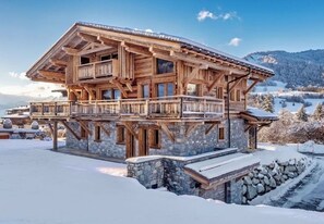 Eine Holzhütte mit Balkon, Steinuntergrund und verschneiter Landschaft im Hintergrund.