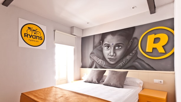 A hotel room with a bed, a nightstand, a wall-mounted artwork, and a logo on the wall. - Ryans Budget (Ibiza Town)