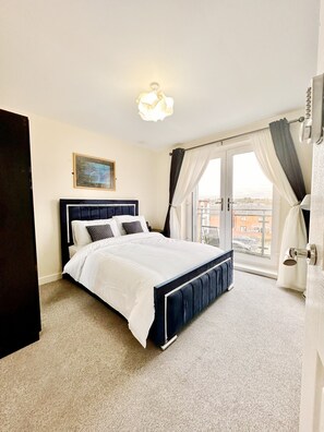 A bedroom with a large bed, a window with curtains, a painting on the wall, and a view of buildings outside. - Luxury 2bed Apartment in Wolverhampton (Wolverhampton)