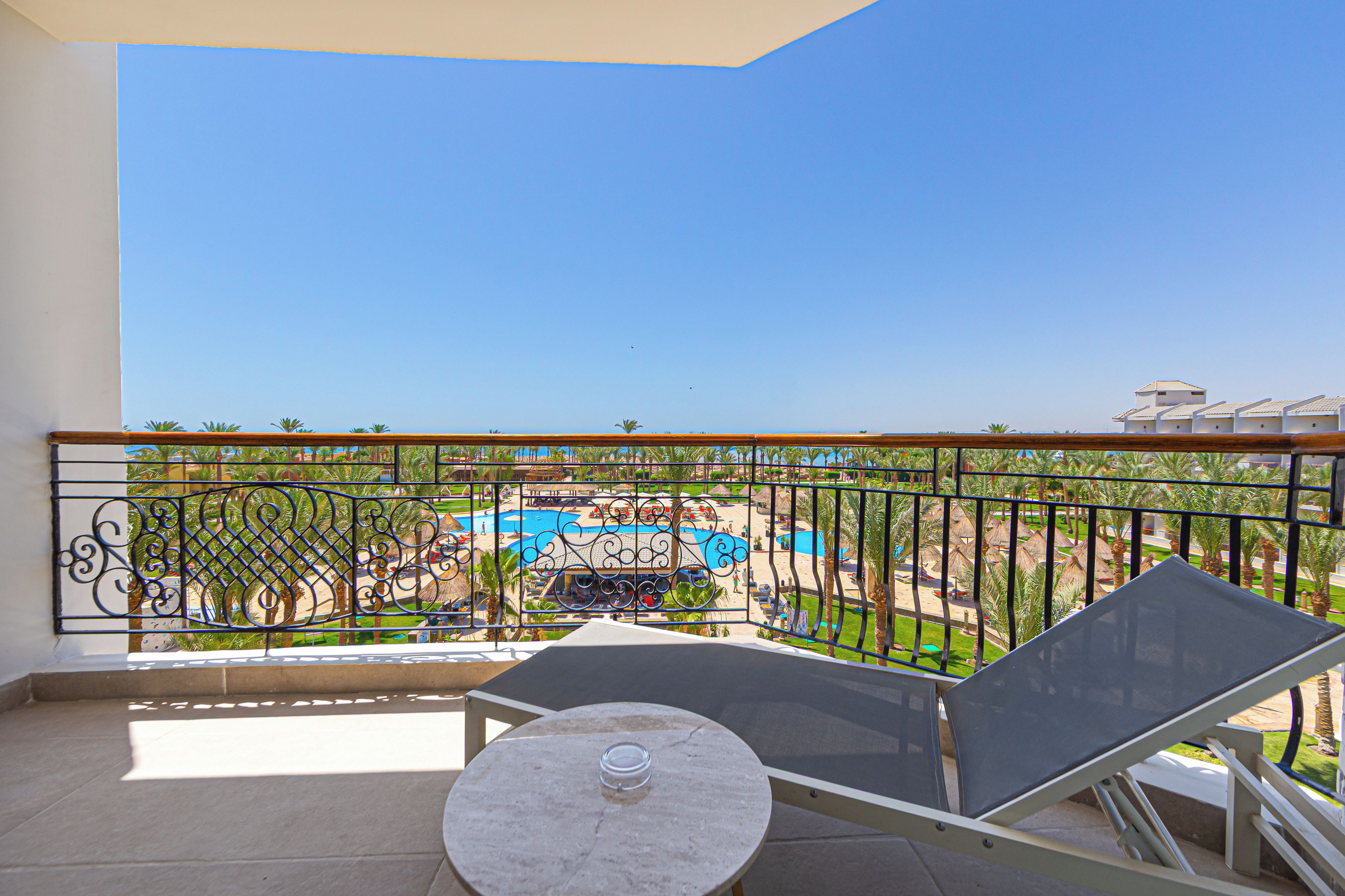 A balcony with a view of a pool area, a lounge chair, and a table.