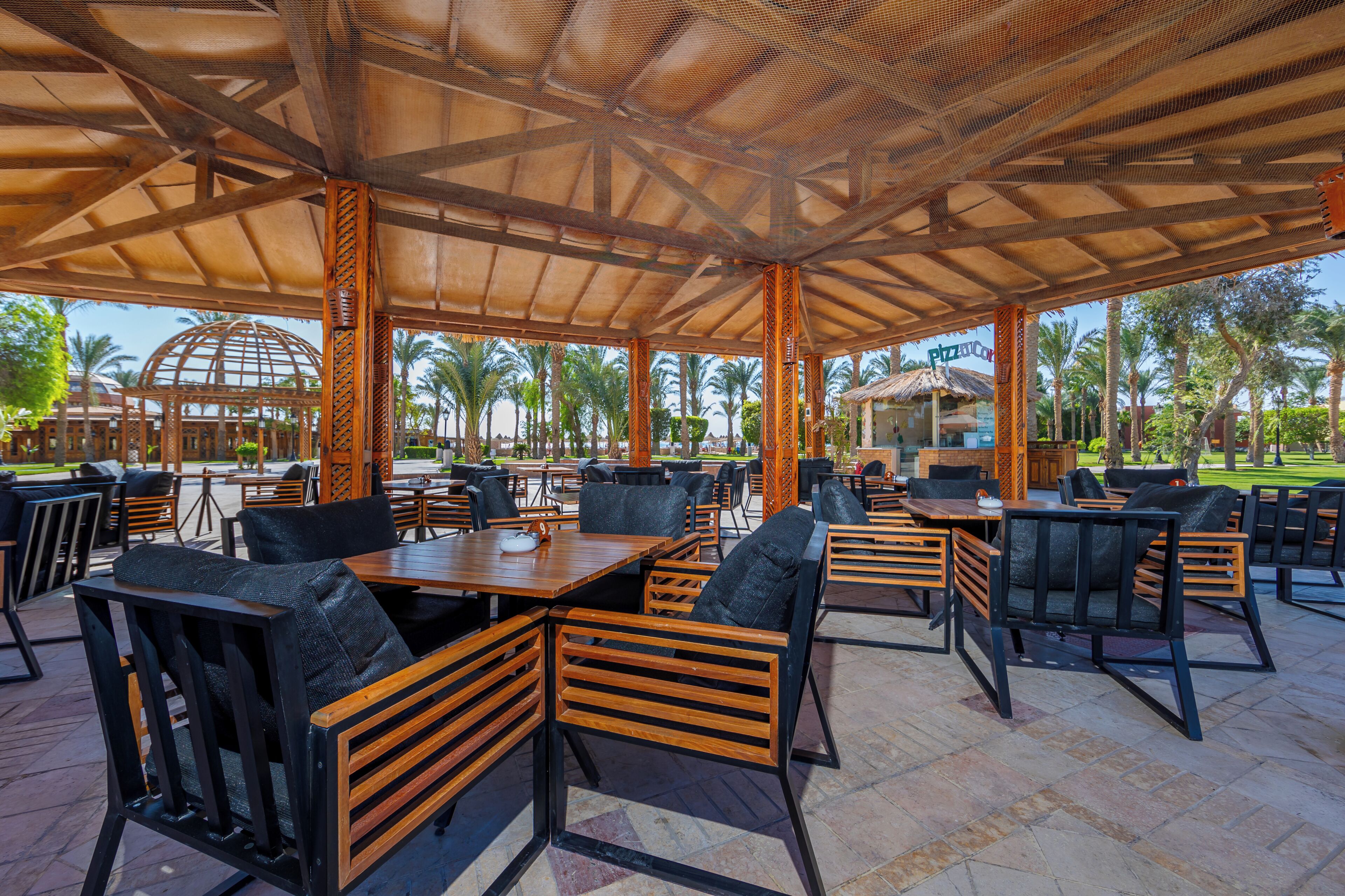 An outdoor patio with wooden tables and chairs, a covered seating area, and a view of palm trees and a dome structure in the background.