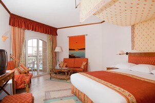 Habitación de hotel con cama, televisor, sofá, sillón, mesa y balcón con cortinas.