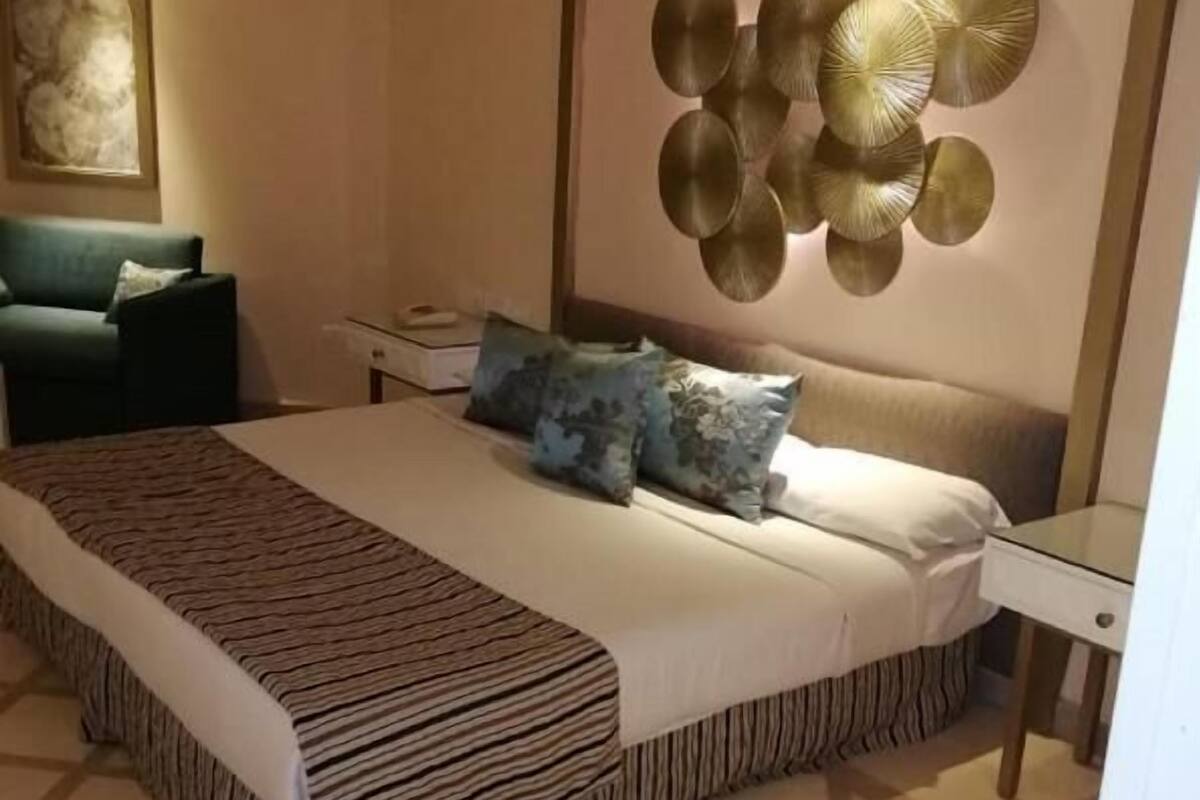 A bedroom with a large bed, a nightstand, a lamp, a chair, and a wall art piece.