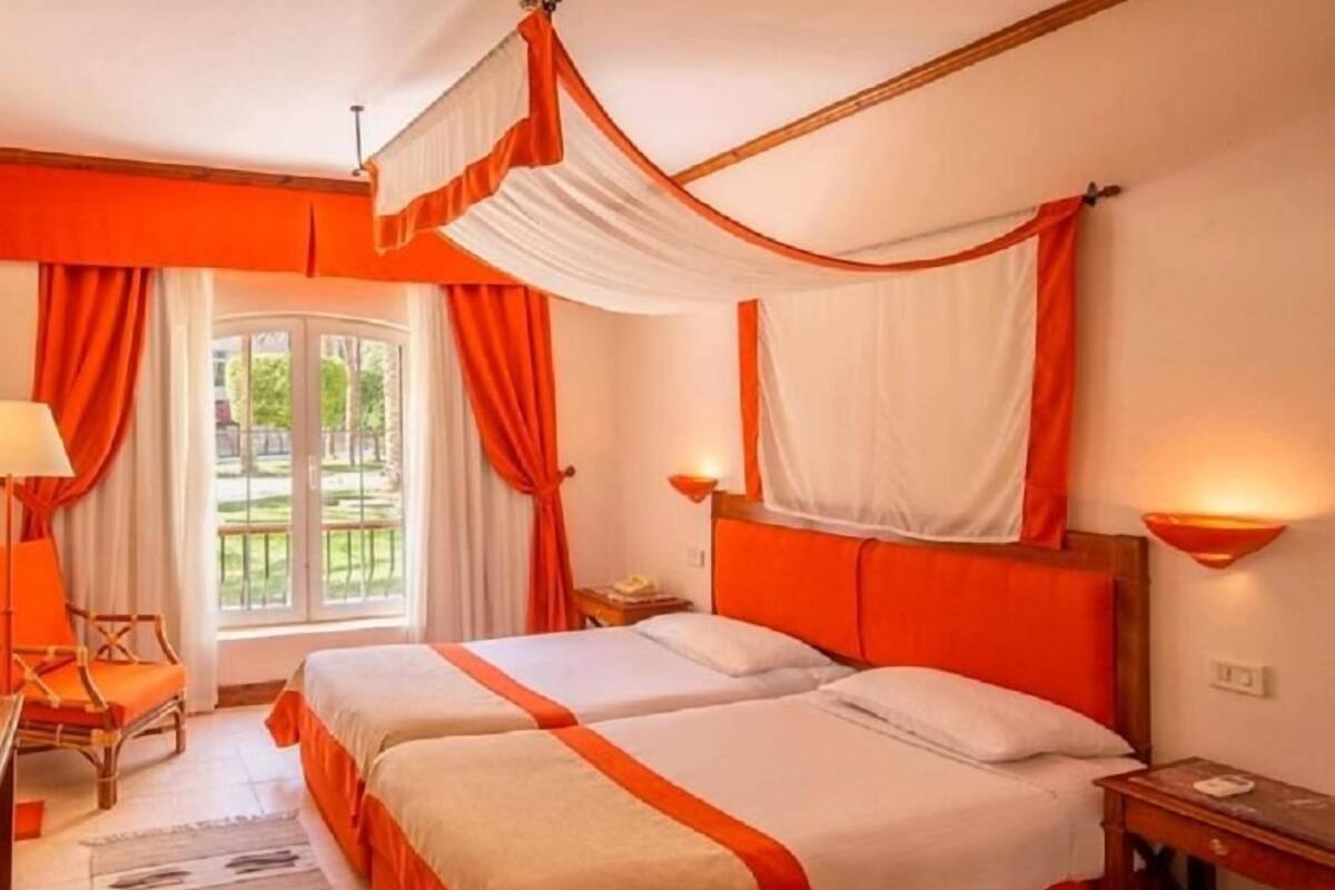 A double bed with a red and white bedspread, a wooden nightstand, a lamp, and a view of the outdoors through a window.