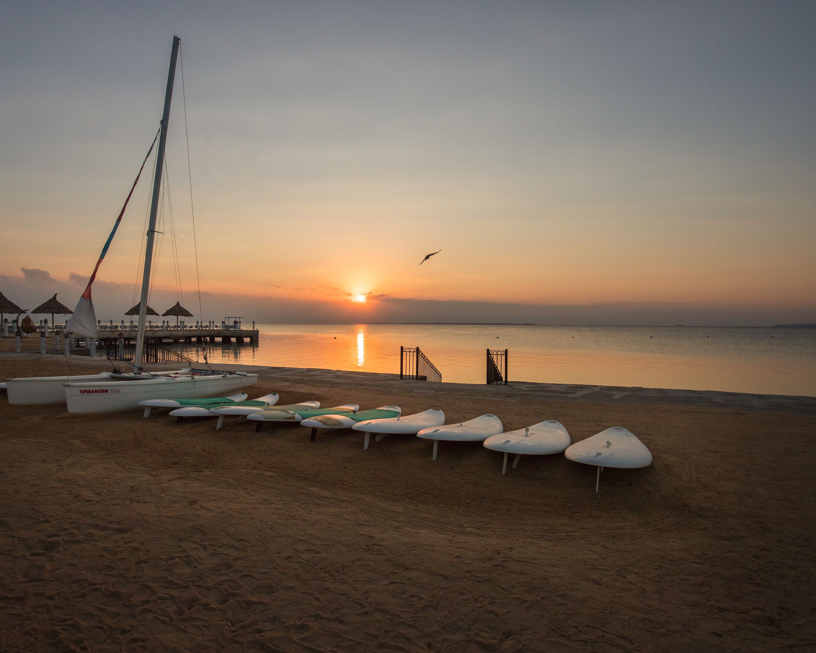 A beach with lounge chairs, a sailboat, and a pier.