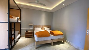 A hotel room with a bunk bed, a single bed with orange pillows, a bedside table with a lamp, and a ceiling fan. - Madpackers Bir  (Baijnath)
