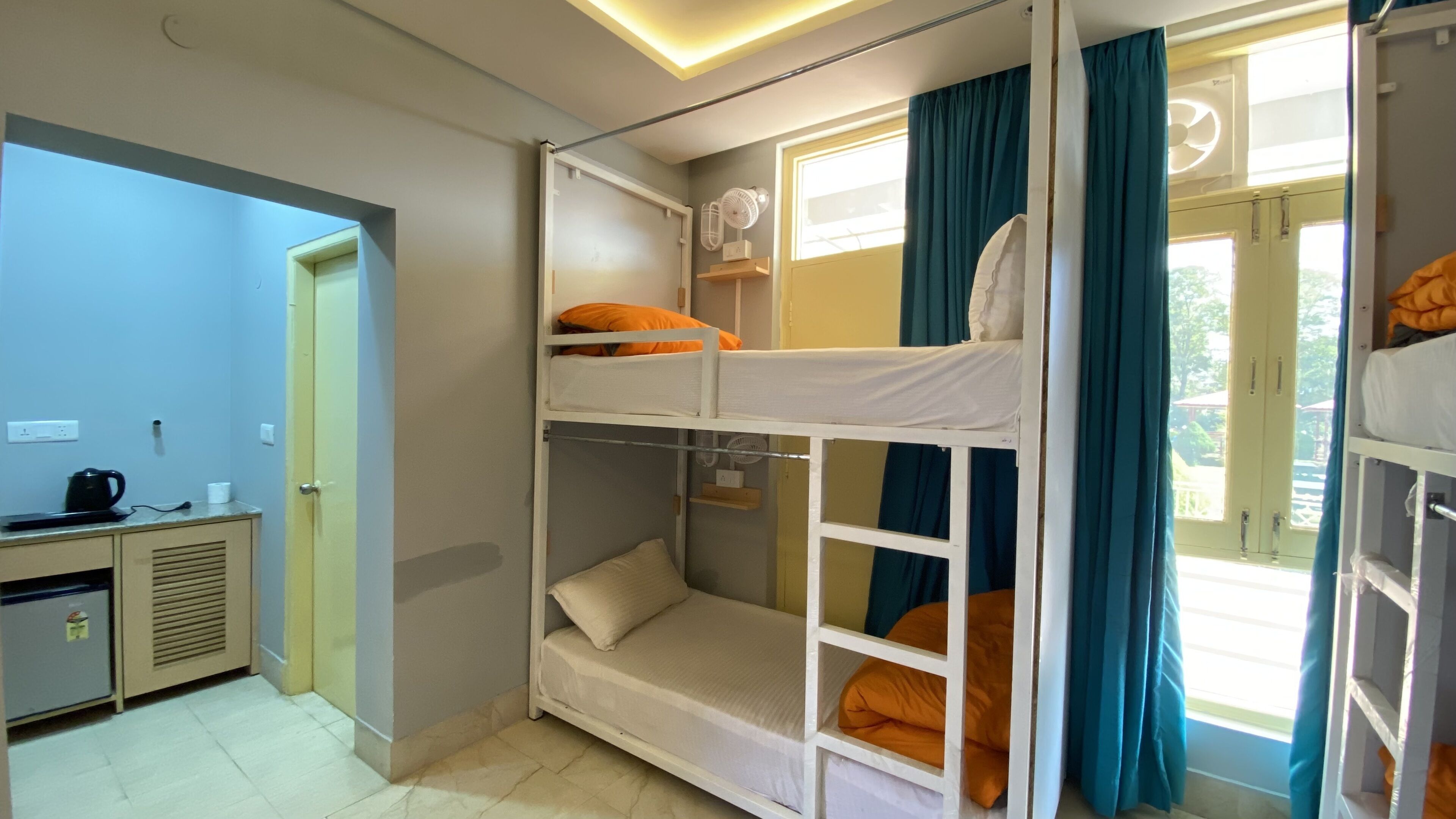 A dormitory room with bunk beds, a desk with a kettle, and a window with curtains.