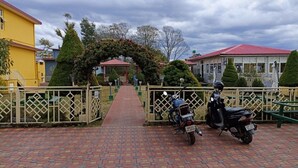 A paved pathway leading to a building with a red roof, surrounded by greenery and a fence. - Madpackers Bir  (Baijnath)