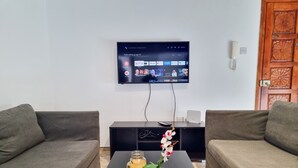 Ein Wohnzimmer mit zwei grauen Sofas, einem schwarzen Couchtisch mit einer Glaskaraffe und Blumen sowie einem flachbildfernseher an der Wand, der verschiedene Streamingdienste zeigt.