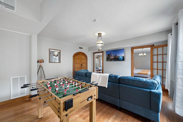 A room with a foosball table, a blue sofa, a wooden door, and a cityscape painting. - Nr Med Center, Museum | WiFi Games | BBQ Fire Pit (Houston)