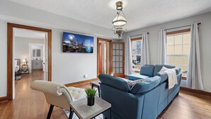A living room with a sofa, a chair, a small table with a plant, a ceiling light fixture, and a cityscape painting on the wall.