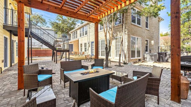 An outdoor patio with wicker furniture and a fire pit, surrounded by brick flooring and wooden pergola.