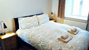 Un letto rifatto con cura, coperto da una trapunta a motivi floreali, due cuscini e due asciugamani arrotolati. Un comodino con una lampada e un cassetto si trova accanto al letto.