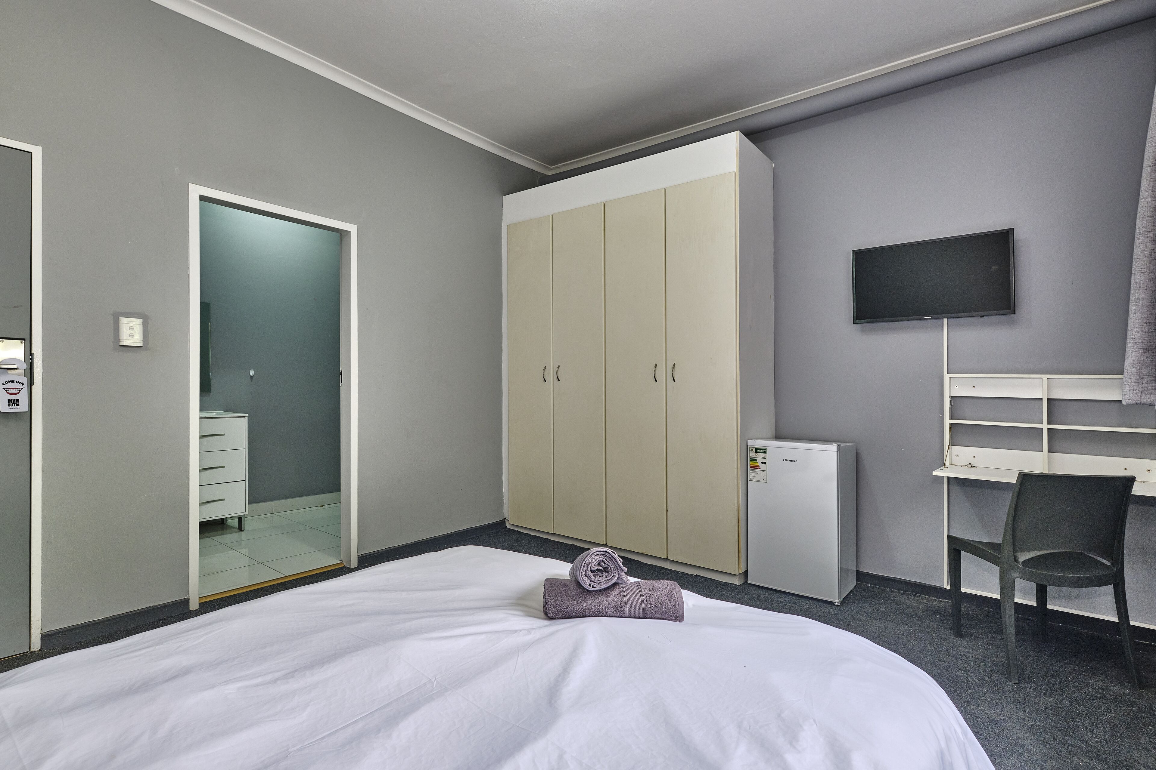 Una camera d'albergo con un letto, una scrivania, una televisione, un armadio e un bagno visibile attraverso una porta aperta.