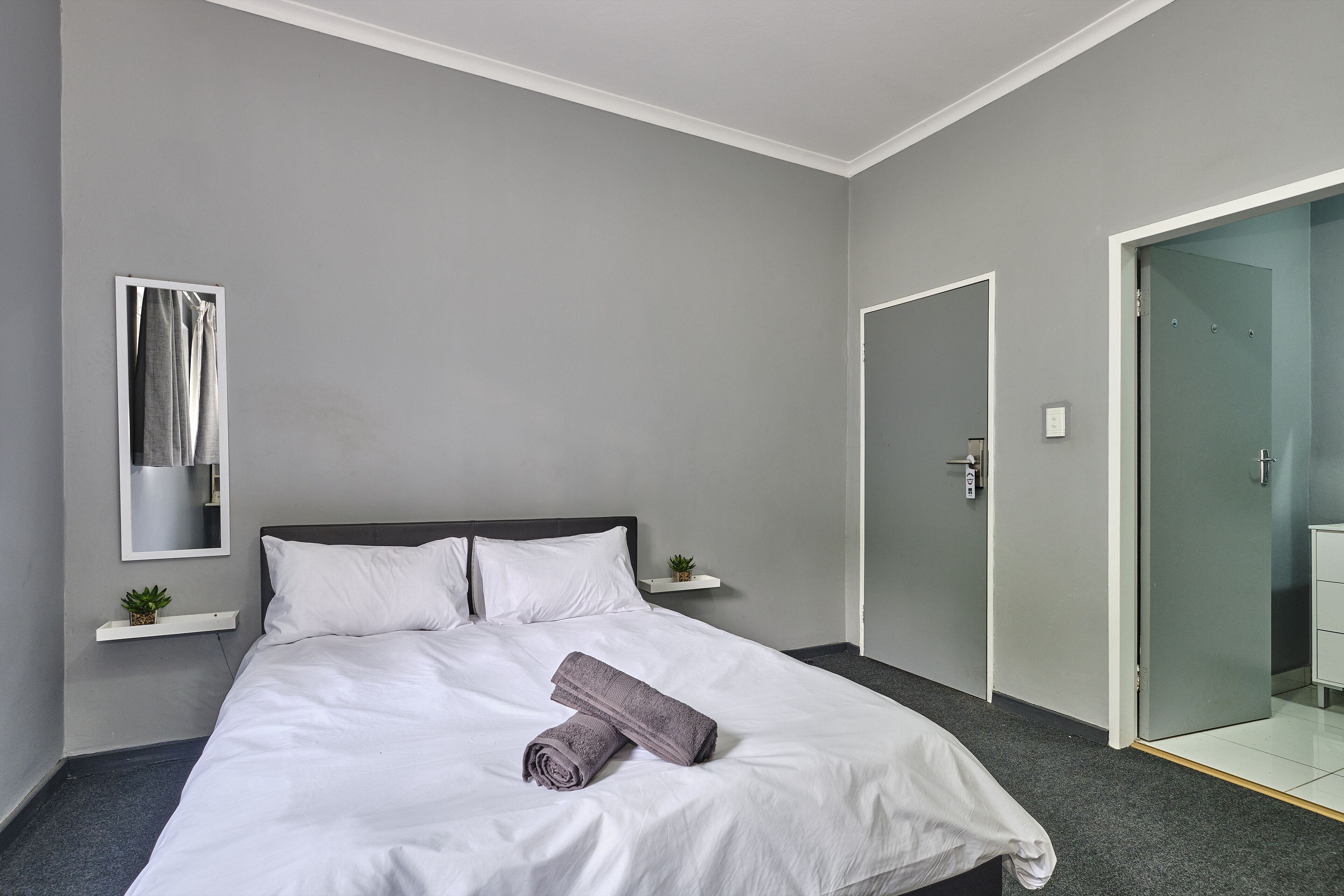 Una cama bien tendida con ropa de cama blanca y una toalla enrollada, una planta pequeña en un estante y un espejo en la pared.