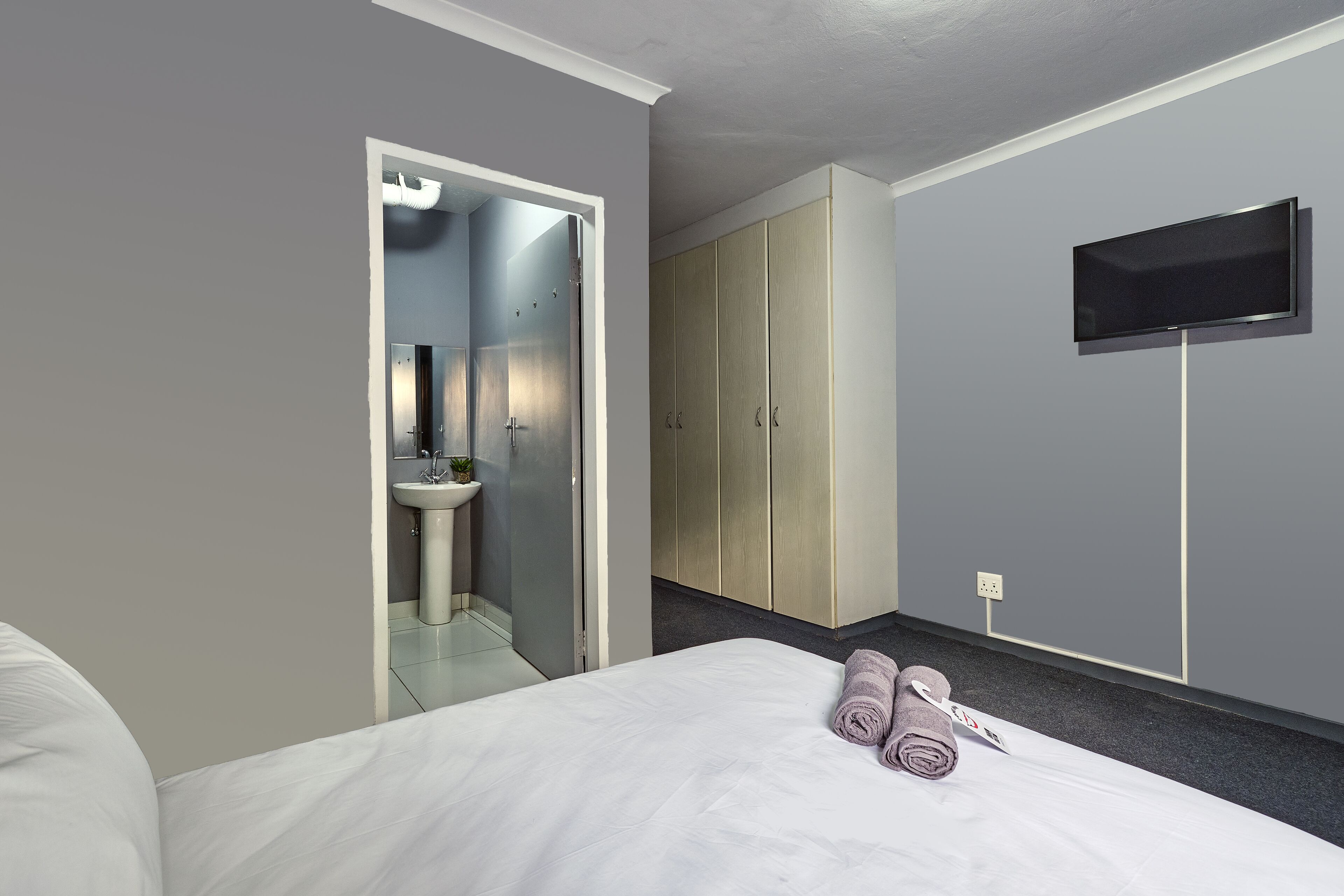 Habitación de hotel con cama, televisor montado en la pared, armario y baño visible a través de una puerta abierta.