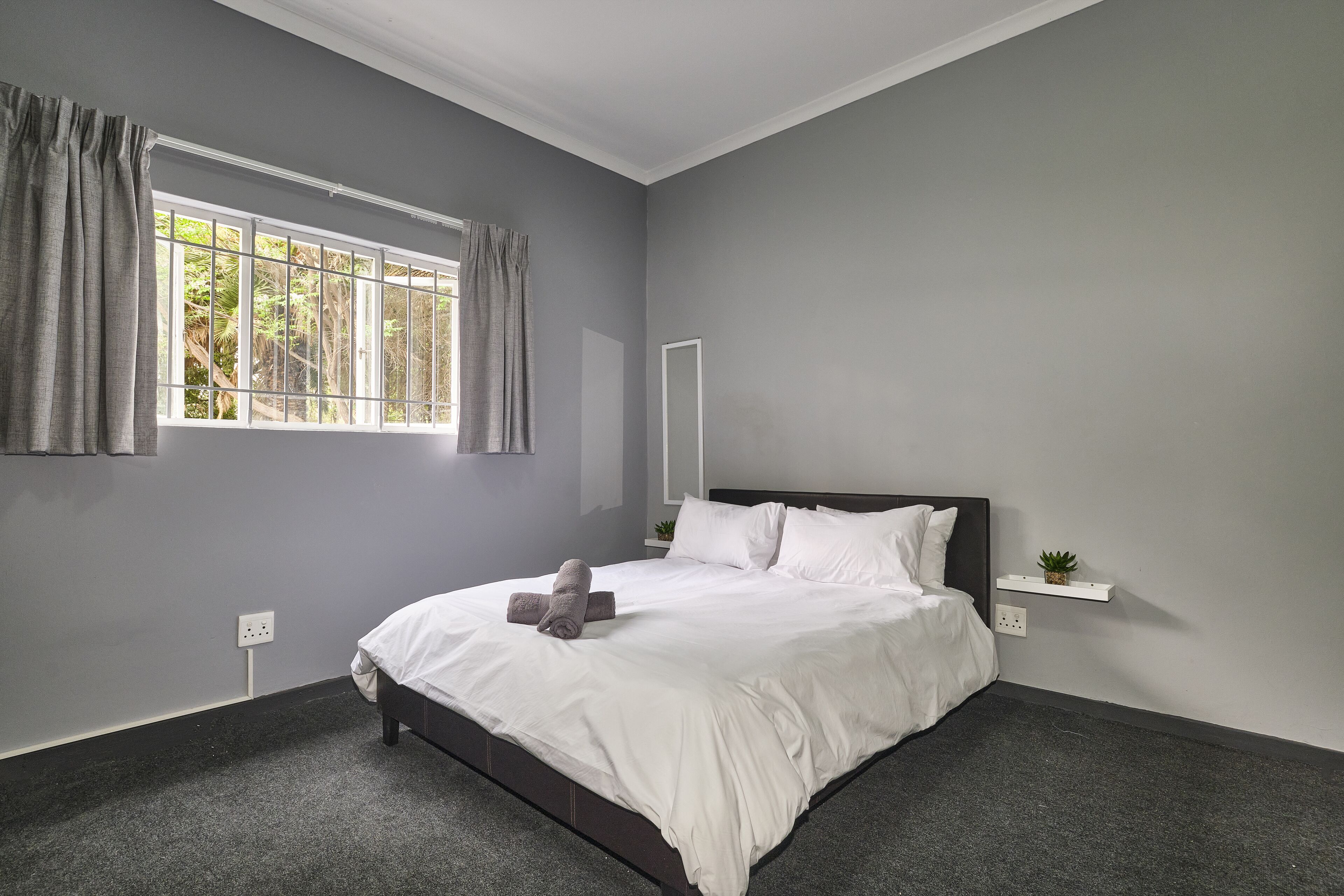 A bedroom with a bed, grey walls, a window with curtains, a bedside plant, and a power outlet.