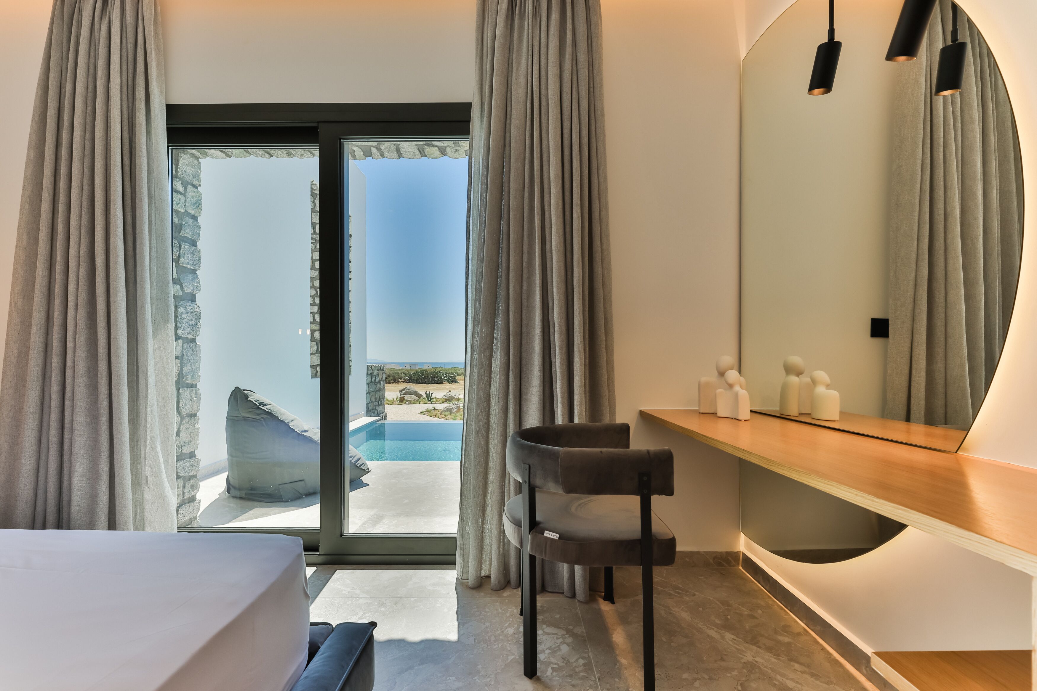 A modern bedroom with a large sliding glass door leading to a pool, a wooden vanity, and a curved mirror.