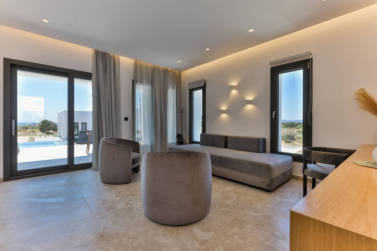 Ein modernes Wohnzimmer mit einem großen grauen Sofa, einem Holztisch und einem grauen Hocker. Zwei große Fenster mit Vorhängen, eine Wandleuchte und Blick auf einen Pool und Grünflächen.
