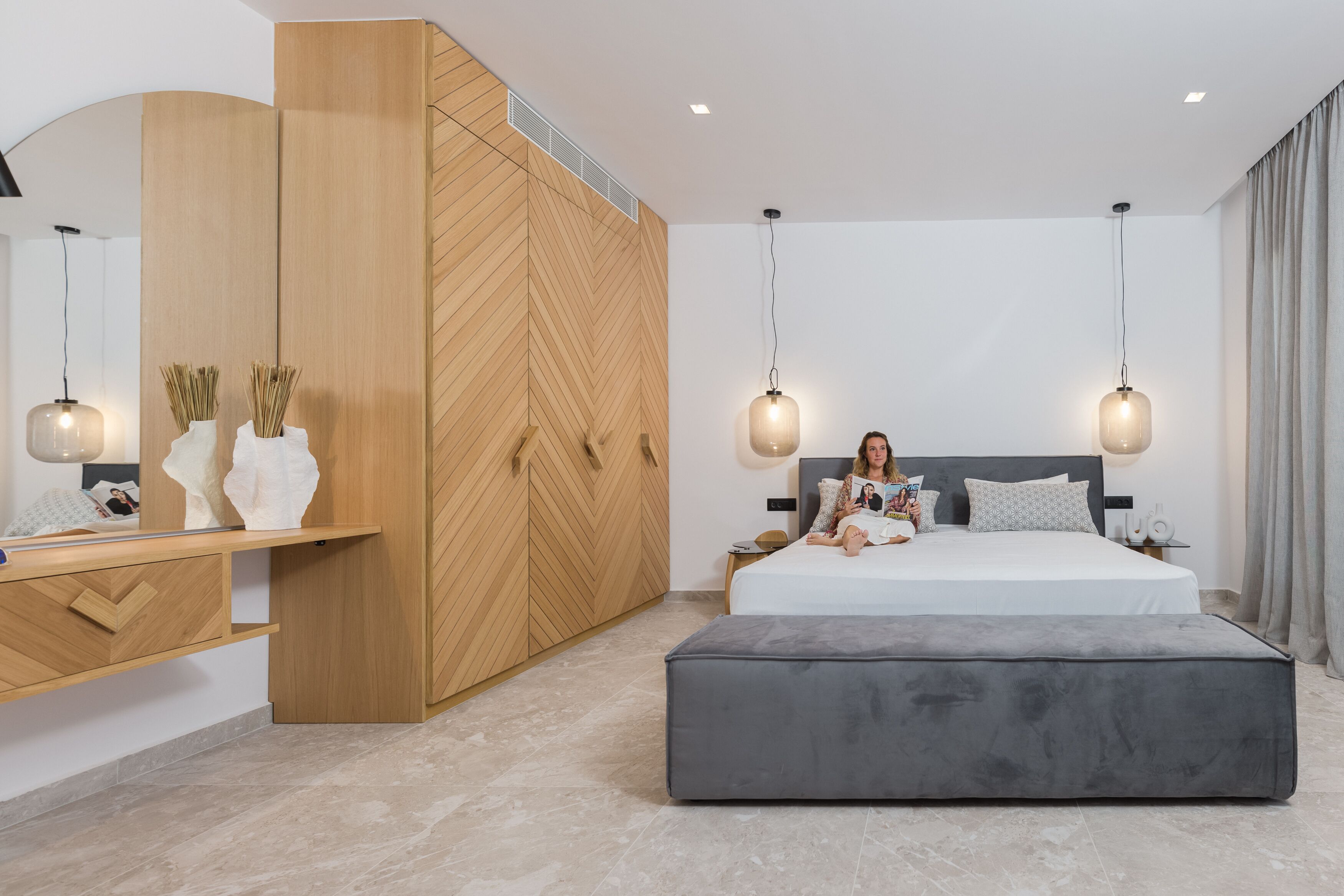 Un dormitorio moderno con una cama grande, un reposapiés gris, un armario de madera e iluminación colgante.