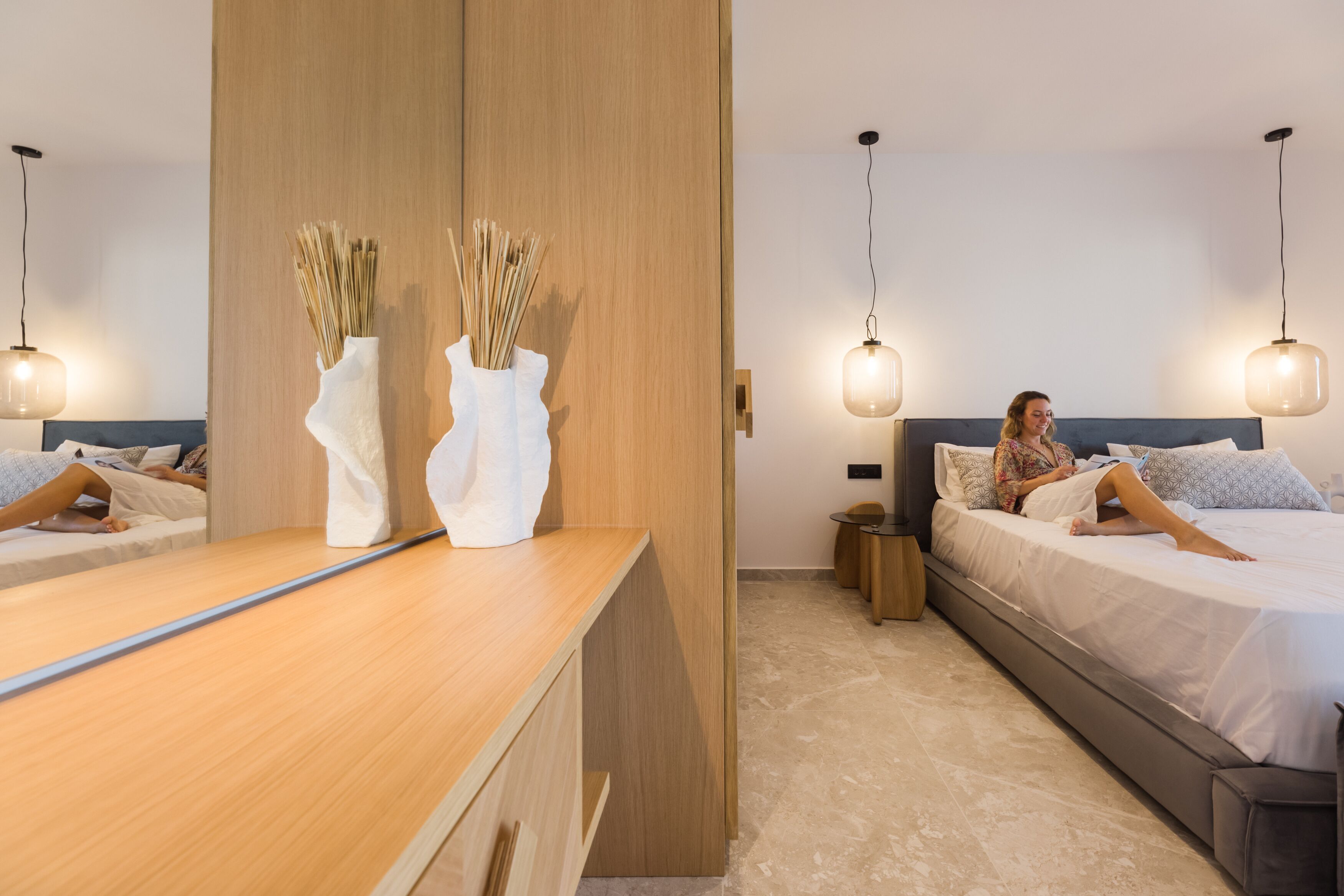 Ein modernes Hotelzimmer mit einem Holzbüro, zwei Nachttischen, einem großen Bett mit grauem Kopfteil und zwei Pendelleuchten.