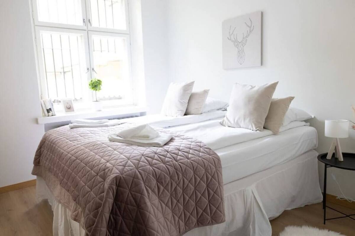 Une chambre à coucher avec un lit, des oreillers, une table de chevet, une lampe, une fenêtre et une œuvre d’art murale.