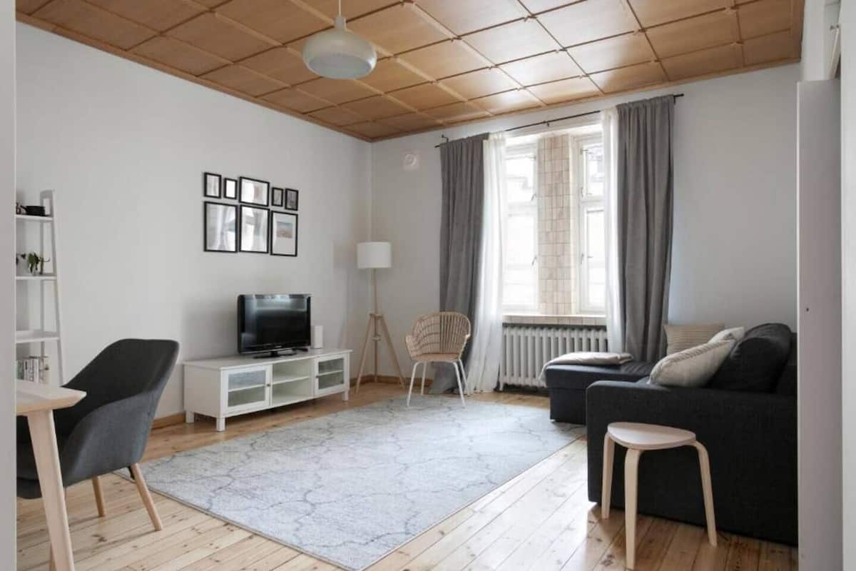 Uma sala de estar moderna com televisão, sofá e poltronas, uma estante branca e uma janela com cortinas.