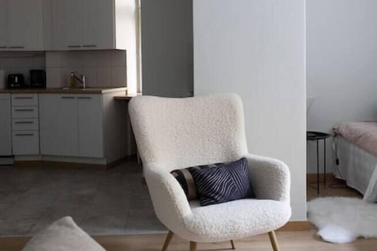 Ein modernes Wohnzimmer mit einem hellen Sessel, einer Couch und einer Küche im Hintergrund.
