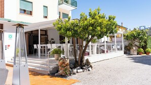 Un bâtiment doté d’un espace extérieur abrité avec des sièges, d’un escalier menant à l’entrée et d’un arbre au premier plan.