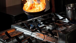 Un chef dans une cuisine professionnelle, avec des flammes, une cuisinière et des ustensiles de cuisine.