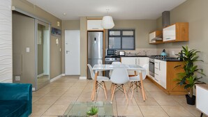 A modern kitchen with white cabinets, a stainless steel refrigerator, and a dining area with a glass table and white chairs. - Knightsbridge 108 (Cape Town)
