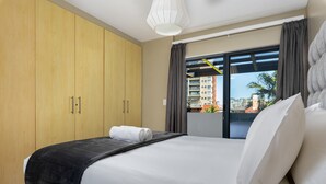 A hotel room with a large bed, a wooden wardrobe, a balcony with a view of buildings and palm trees. - Knightsbridge 108 (Cape Town)