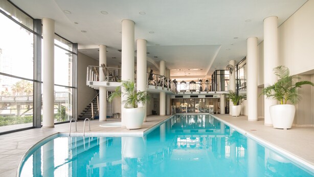 An indoor swimming pool with clear blue water, surrounded by large white columns and potted plants. - Knightsbridge 108 (Cape Town)