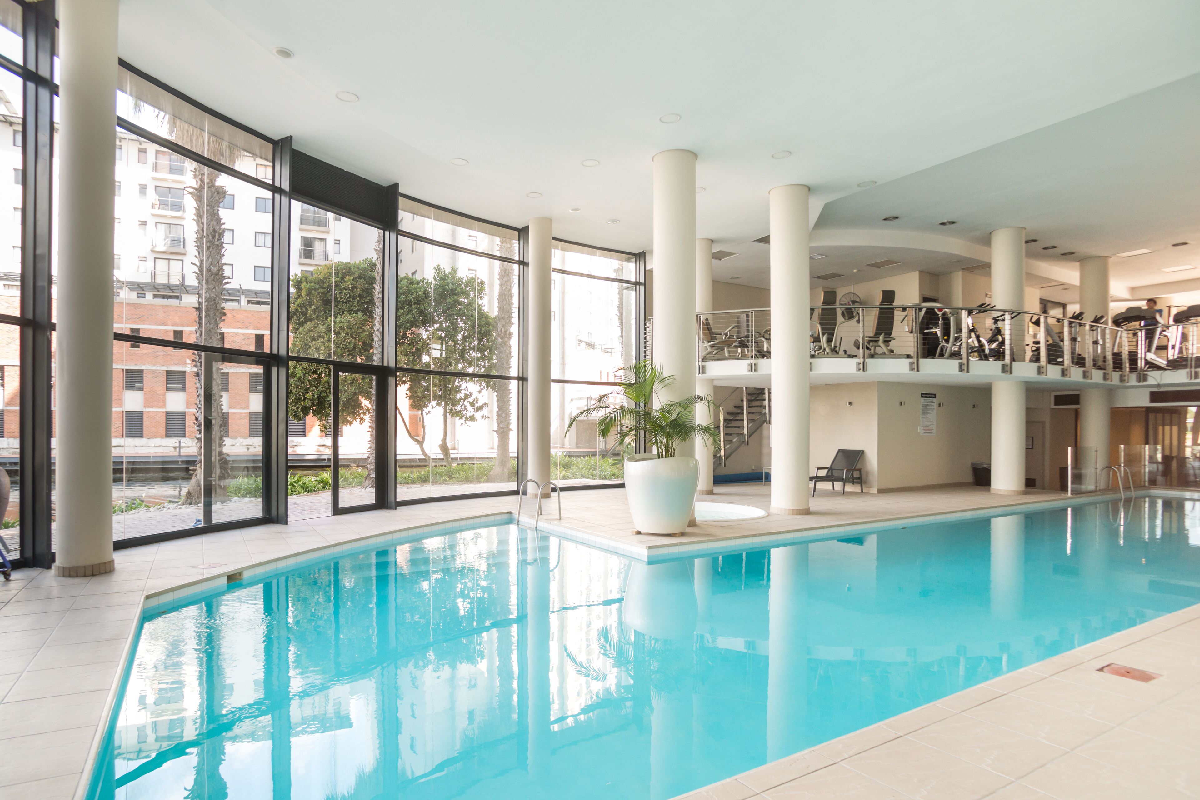 An indoor swimming pool with clear glass walls, a potted plant, and a staircase leading to an upper level.