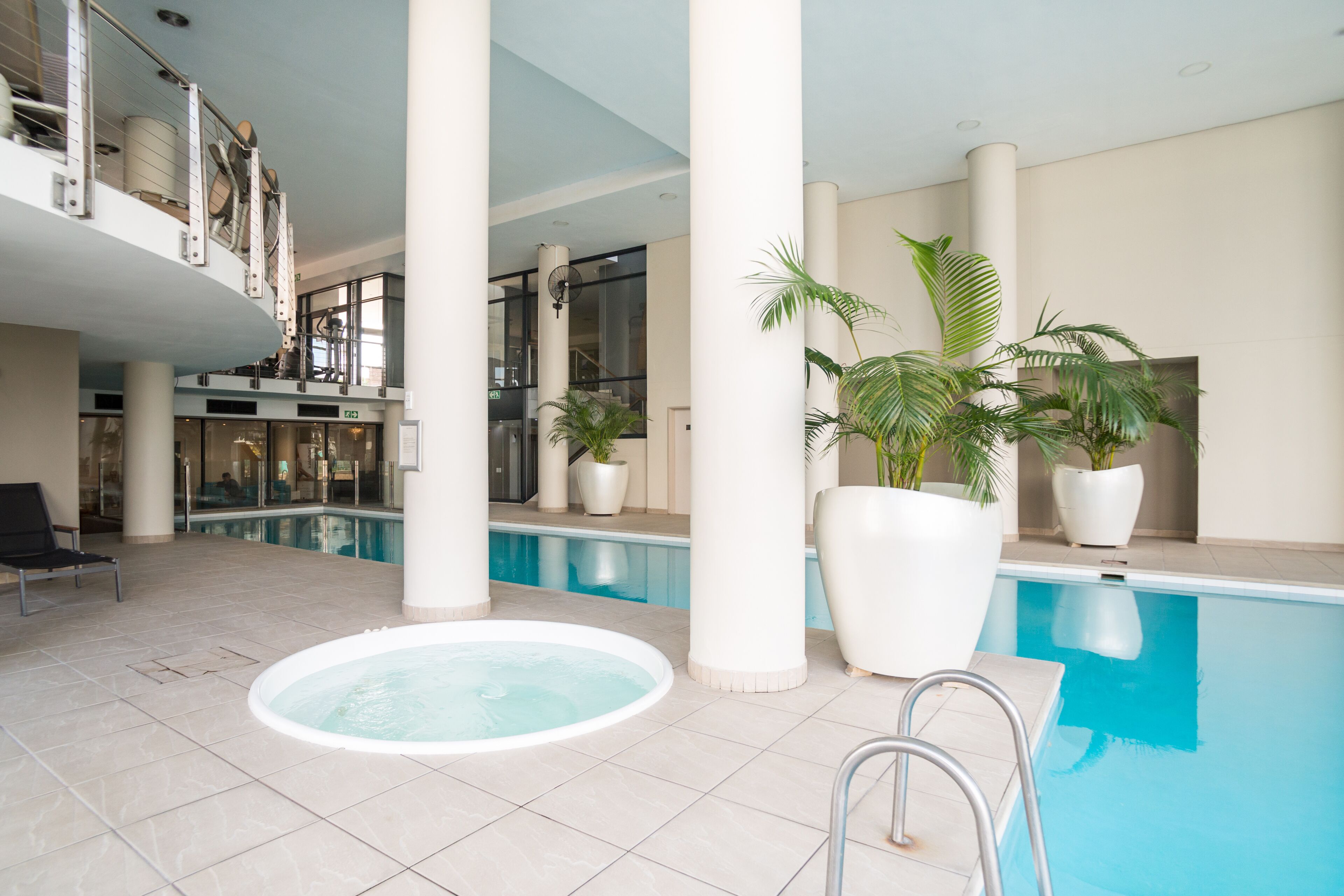 Une piscine intérieure dotée d’un jacuzzi, entourée de grandes colonnes blanches, de plantes en pot et d’un fauteuil.