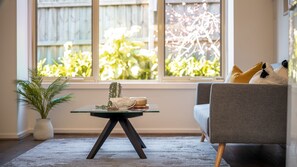 A living room with a glass coffee table, a sofa, a potted plant, and a view of greenery through large windows. - Sunshine Retreat Villa 2 (Cowes)
