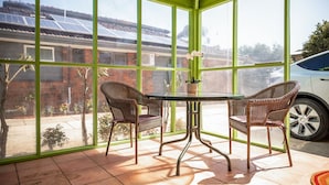 Une véranda avec une table en verre et des chaises en osier, une plante en pot, et une vue sur un bâtiment en briques et des panneaux solaires.