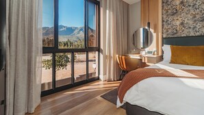 A hotel room with a large window offering a mountain view, a bed with a brown blanket, a desk with a chair, and a modern wall design. - The Hawthorn Boutique Hotel (George)