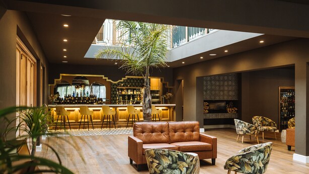 A spacious lounge area with a leather sofa, patterned armchairs, a central tree, and a bar with shelves of bottles. - The Hawthorn Boutique Hotel (George)