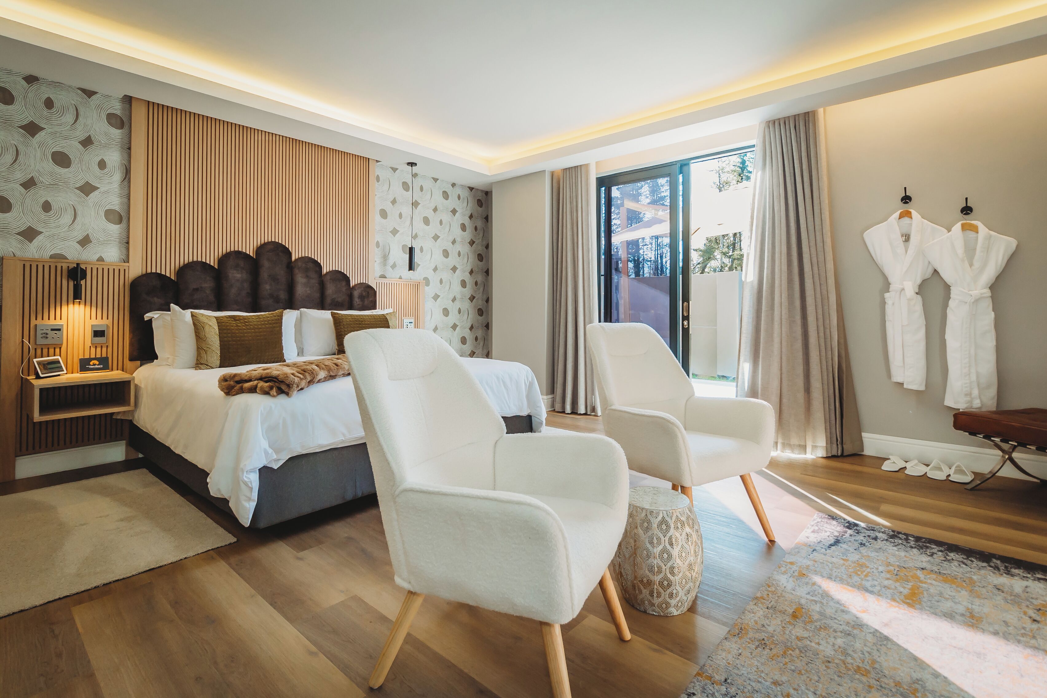 Quarto de hotel moderno com uma cama grande, duas poltronas brancas, uma mesinha de apoio, uma janela ampla com cortinas e dois roupões brancos pendurados na parede.