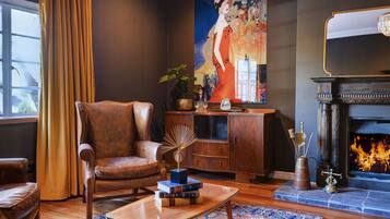 Uma sala de estar com uma poltrona de couro, uma mesa de centro de madeira com livros, uma lareira e um grande quadro abstrato na parede.