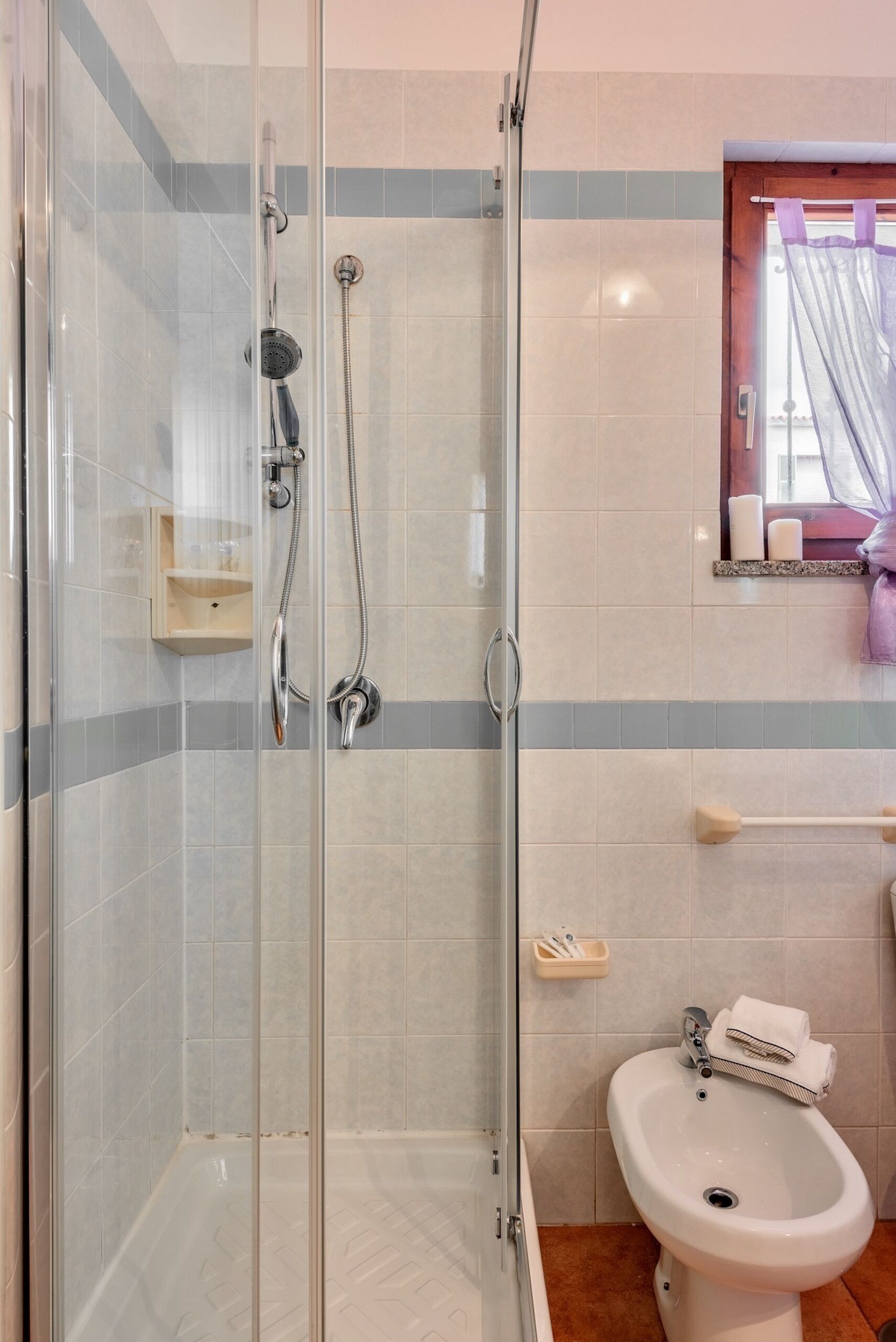 A bathroom with a glass-enclosed shower, a white sink, and a window with a purple curtain.