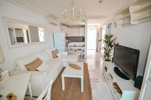 A modern living room with a white sofa, a coffee table, a TV, and a kitchen in the background.
